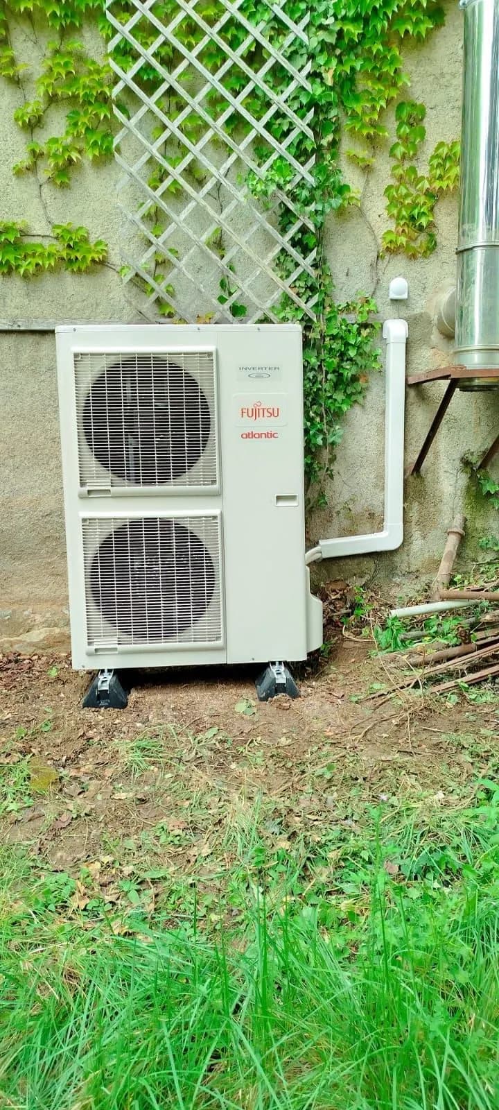 Unité extérieure de pompe à chaleur à double ventilateur, posée sur plots contre un mur de vigne vierge